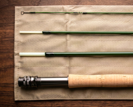 A disassembled fly fishing rod laid out on a beige fabric roll over a dark wood surface. The rod sections show a green shaft, white plugs, and a cork handle with a metal reel seat.