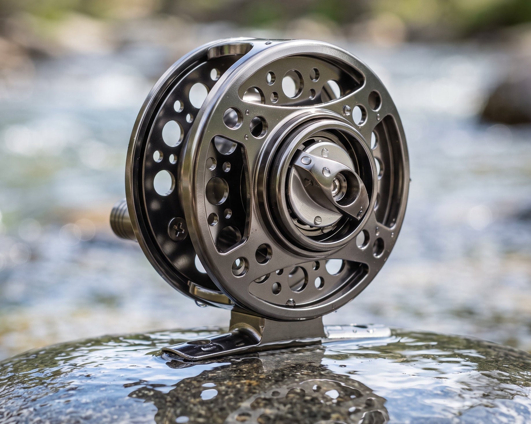 A close-up of a metallic fly fishing reel mounted on a rock in a flowing stream, with blurred water and trees in the background.