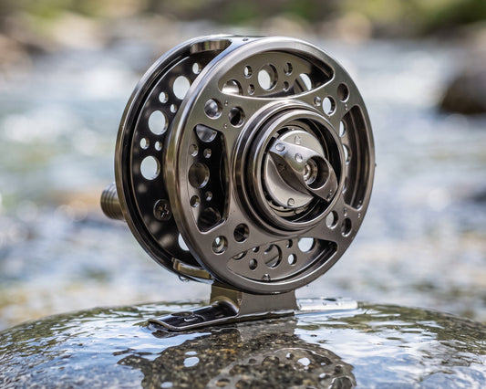 A close-up of a metallic fly fishing reel mounted on a rock in a flowing stream, with blurred water and trees in the background.