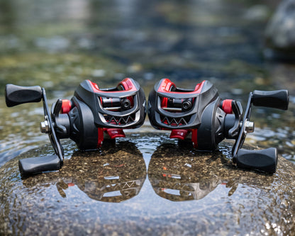Two black and red baitcasting fishing reels resting on a wet rock in shallow water, with a blurred natural outdoor background.