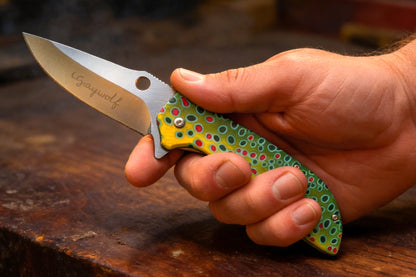 Hand holding a colorful folding knife with 'Graywolf' branding on a wooden surface.